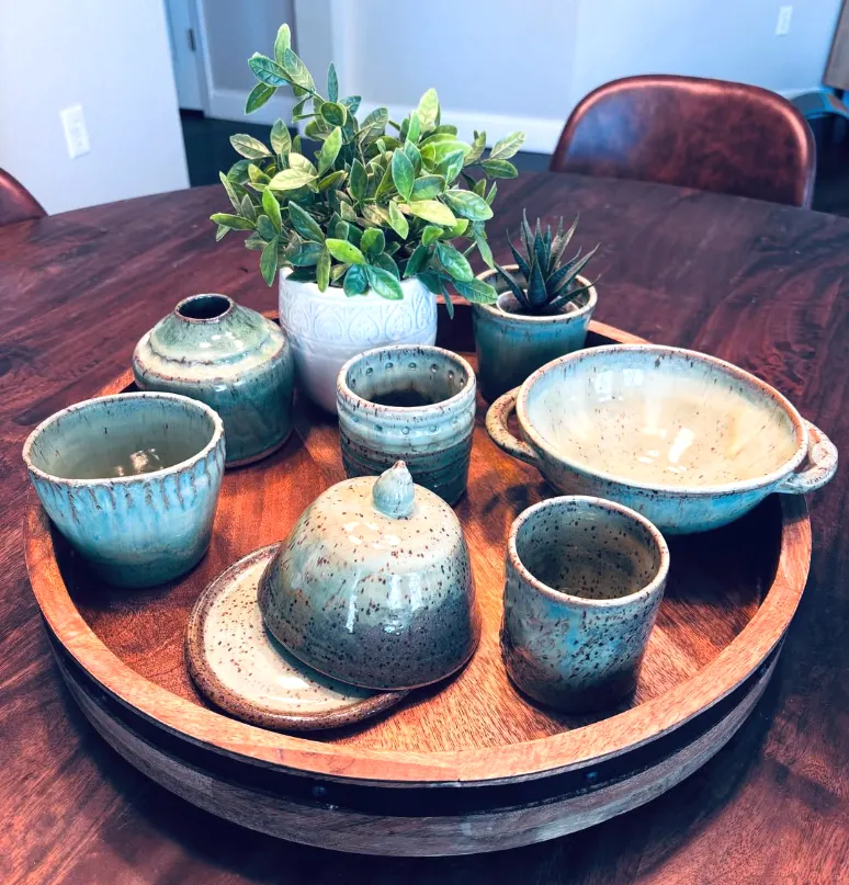 A tray of finished pieces in blue and green glazes: beakers, a lidded butter dish, a vase and a handled bowl.
